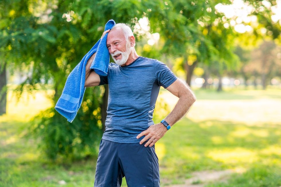 Idoso com cabelos brancos e barba, vestindo roupas de ginástica azuis, faz uma pausa enquanto se exercita ao ar livre pela manhã